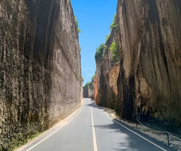 Smooth asphalt road carved directly through towering, 20-meter-high sheer limestone canyon walls at Tanah Barak Cliff